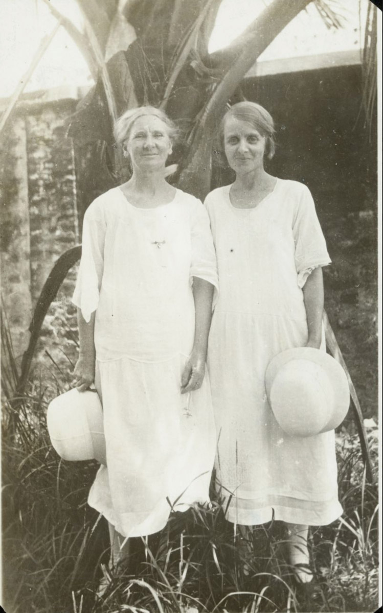 black and white outdoor portrait of two people in long white dresses, each holding a hat and standing in front of a tree